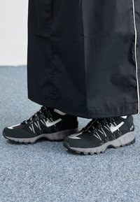 Černé sportovní boty s texturovaným síťovaným svrškem, stříbrnými akcenty a robustní gumovou podešví. Kombinovány s dlouhým černým oděvem.