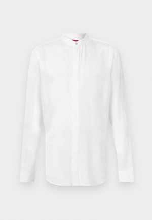 Chemise blanche à manches longues avec col officier et patte de boutonnage cachée, présentée sur un fond clair uni.