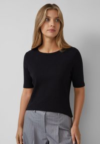 T-shirt nera a coste con maniche corte e scollo rotondo, indossata con pantaloni grigi a righe sottili. Il tessuto sembra morbido e aderente.