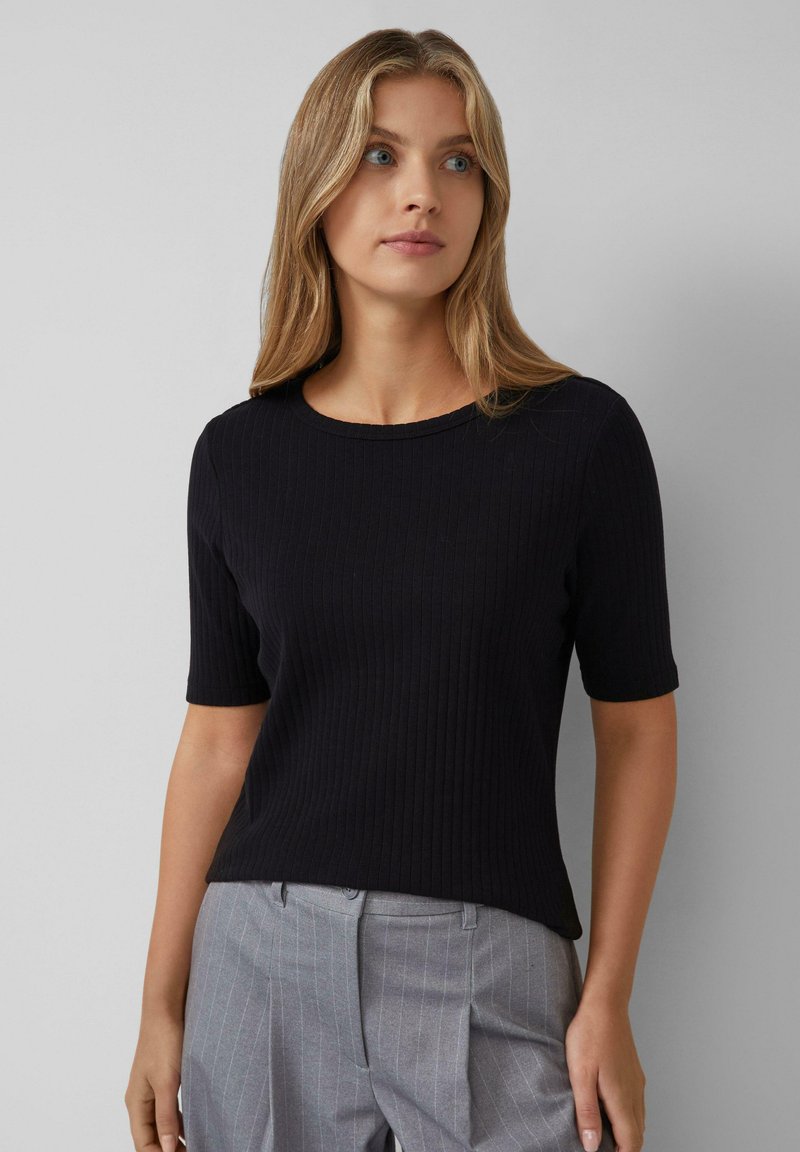 T-shirt nera a coste con maniche corte e scollo rotondo, indossata con pantaloni grigi a righe sottili. Il tessuto sembra morbido e aderente.