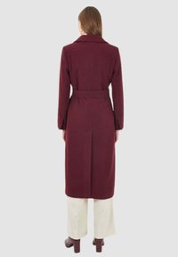 Cappotto color mogano con vita cintata, colletto strutturato e maniche lunghe. Presenta una silhouette dritta e tessuto texturizzato.