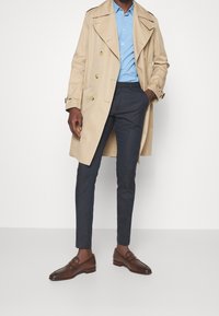 Ljus beigefärgad trenchcoat, ljusblå skjorta, marinblå byxor och mörkbruna läderskor. Kappan har dubbelknappning och axelremmar.