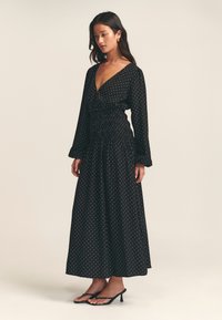 Black, polka dot maxi dress with a deep V-neck, long sleeves, and a smocked waist. Designed with a flowy silhouette and lightweight fabric.