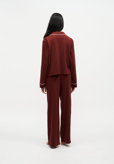 Femme aux longs cheveux foncés, debout pieds nus de dos, portant un haut bordeaux à manches longues et un pantalon large avec une bordure blanche aux poignets et au col.