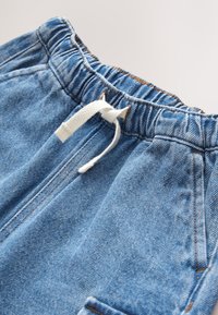 Blå denim-bukselinning med elastik og hvid snor bundet i en simpel knude, synlige syninger og kanter på forlommerne.