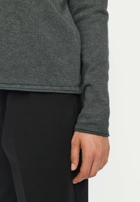 Maglietta a maniche lunghe grigia scura con una texture liscia, orlo arrotondato e pantaloni neri a contrasto. La manica presenta un dettaglio con polsino cucito.