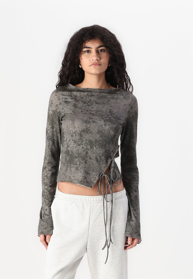 Grijs cropped top met lange mouwen en een gestructureerd patroon, zijsnoerdetails en een ongelijkmatige zoom. Gecombineerd met lichtgrijze broek.
