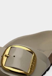 Μπεζ σανδάλι με γυαλιστερό φινίρισμα, με φαρδιά λουρίδα και μεγάλο χρυσό αγκράφα. Η σόλα έχει υφή, προσφέροντας άνεση και σταθερότητα.