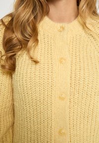 Dzeltena adīta kardigāna ar teksturētu rakstu, ar apaļu kakla izgriezumu un piecām apaļām pogām priekšpusē. Mīksts, ērts materiāls.