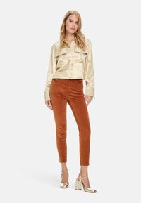 Satinbluse in Gold mit zwei Taschen, kombiniert mit einer rostfarbene Cordhosen und metallischen Blockabsatzsandalen mit Riemchen.