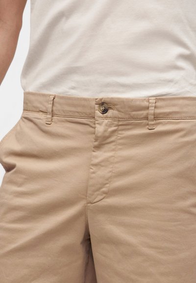 Pantalon en coton beige avec fermeture par bouton et fermeture éclair, porté avec un haut crème rentré.