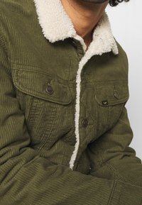 Giacca in corduroy verde oliva con colletto in shearling crema, dotata di due tasche a patta e bottoni metallici. Tessuto texturizzato visibile.