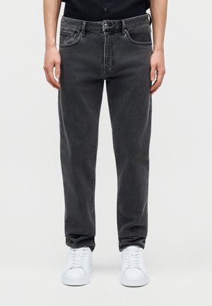 Inimene, kes kannab musta vaba lõikega teksapükse ja valgeid vabaajajalatseid, seisab ühevärvilise heleda tausta ees.