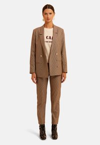 Blazer de cuadros marrón de doble botonadura combinado con pantalones ajustados a juego y una sudadera gráfica de color claro. Botines negros brillantes hasta el tobillo.