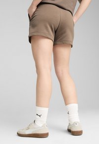 Shorts en coton marron avec revers, associés à des chaussettes blanches à côtes portant un logo noir, et des baskets gris clair avec des semelles marron.