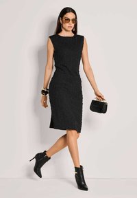 Schwarzes, tailliertes Kleid mit strukturierter Musterung, ärmelloses Design, knielang, kombiniert mit Ankle Boots und einer kleinen schwarzen Tasche.