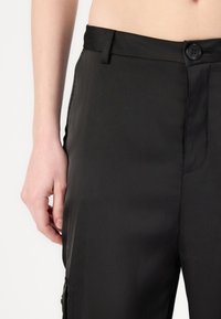 Kerged mustad püksid, mis on valmistatud kerge kangaga, esise nupuga sulgemine, küljetaskud ja sile tekstuur.