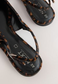 Sandali con stampa leopardata e cinturini in pelle nera, con suola piatta e superficie testurizzata simile a pelliccia. Logo dorato sulla soletta.