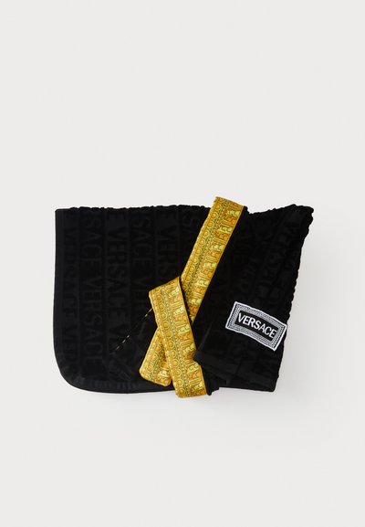 Prosop negru pliat cu textul „Versace” în relief, având o panglică aurie cu model și o etichetă albă cu logo-ul Versace într-un colț.