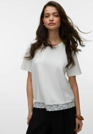 Pikade tumedate juustega naine kannab valget lühikeste varrukatega silmapaistva pitsäärisega topi ja musta pükse, seisab tavalise heleda tausta ees.