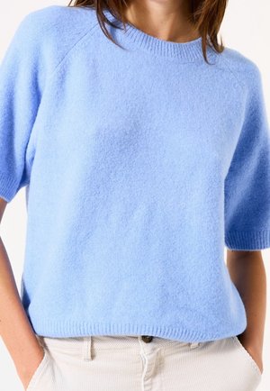 Person trägt einen hellblauen, kurzärmeligen, flauschigen Pullover und beige Hose mit den Händen in den Taschen vor weißem Hintergrund.