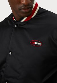 Black bomber jacket with a ribbed red and white collar, featuring a circular embroidered logo in red, black, and white on the chest.