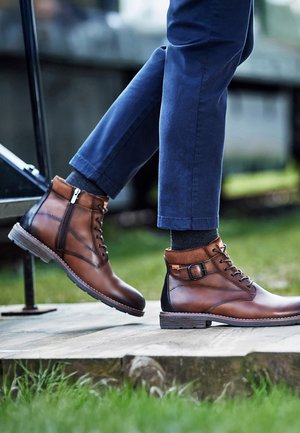 Stivaletti marroni in pelle con lacci, zip laterale e fibbia, indossati con pantaloni blu che camminano all'aperto su una superficie in legno.