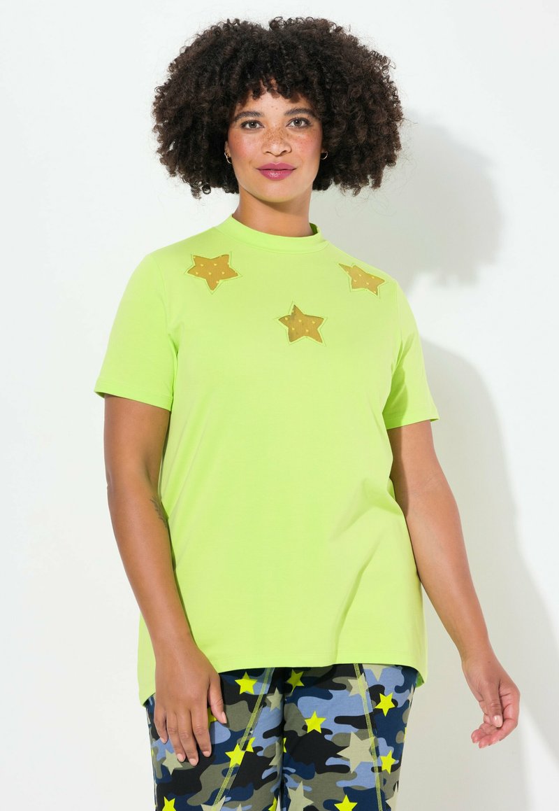 Limegrøn kortærmet t-shirt med høj halsudskæring, der har tre gule stjerne-applikationer på forsiden. Sammen med mønstrede camopants.