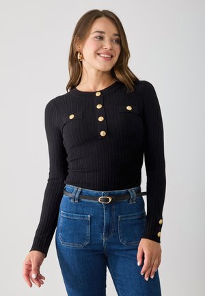 Top lungo a maniche nere a coste con bottoni dorati e dettagli tasca, abbinato a jeans in denim blu a vita alta e una cintura nera.