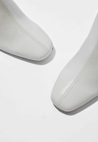 Botas de tobillo de cuero blanco con punta afilada, que presentan costuras visibles y una textura suave sobre un fondo liso.
