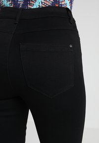 Jeans de denim negro con un diseño ajustado. Presenta un único bolsillo trasero con un detalle de botón, textura suave y cintura de altura media.