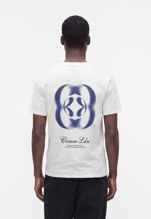 Uomo che indossa una maglietta bianca con un disegno astratto circolare blu e la scritta Closure Ldn sulla schiena, in piedi davanti a uno sfondo bianco uniforme.