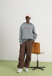 Grauer Kapuzenpullover mit einem Logo, braune Cargo-Hosen und weiße Sneakers. Die Mütze ist hellrosa. Der Hintergrund zeigt eine helle Farbe und einen Holzstuhl.
