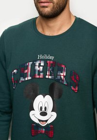 Zelená mikina s vyšitým nápisem „Holiday CHEERS“ v károvaném vzoru a velkou potištěnou hlavou Mickey Mouse s detailem motýlka.