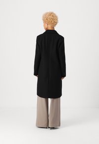 Cappotto nero in lana con colletto a risvolto e maniche lunghe, caratterizzato da una silhouette dritta. Indossato sopra pantaloni beige a gamba larga.