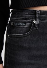 Jeans de mezclilla negra con un lavado oscuro, bolsillo delantero con un botón plateado y etiqueta del logo de Calvin Klein en la cintura.