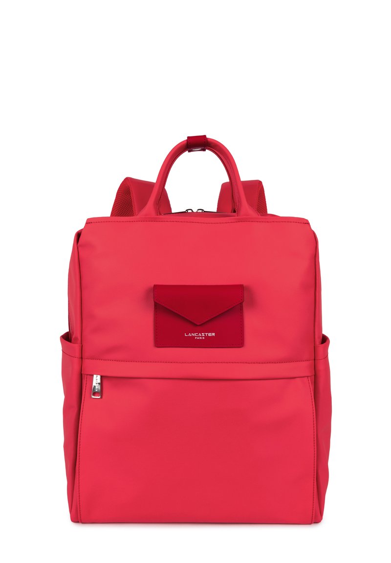 Sac à dos en nylon rouge avec une poche à rabat à l'avant, une poignée supérieure, des bretelles rembourrées et une fermeture éclair. Texture lisse et design minimaliste.
