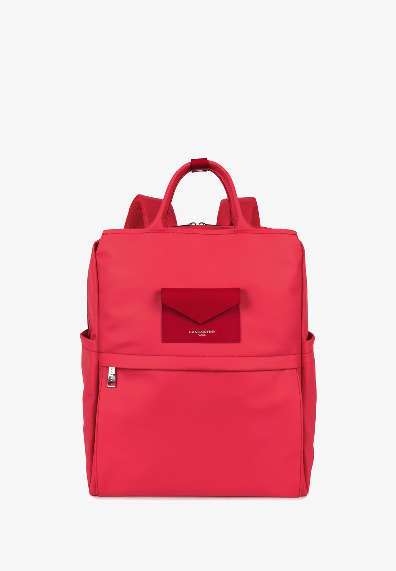 Sac à dos en nylon rouge avec une poche à rabat à l'avant, une poignée supérieure, des bretelles rembourrées et une fermeture éclair. Texture lisse et design minimaliste.