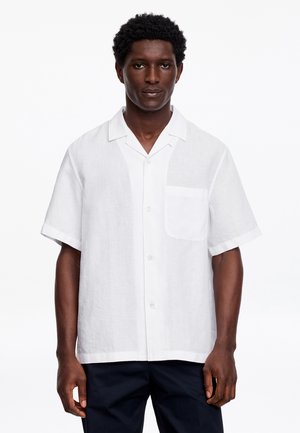 Homme portant une chemise blanche à manches courtes avec une poche poitrine et un pantalon noir, debout devant un fond blanc uni.