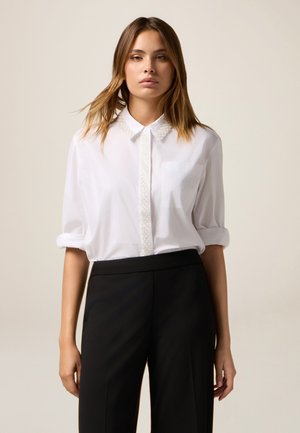 Woman wearing a white buttoned shirt with pearl details on collar and placket, paired with black high-waisted trousers, standing against plain background.