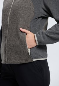 Harmaa vetoketjullinen takki, jossa on teksturoitu materiaali yläosassa, edessä vetoketjullinen tasku ja kontrastiväriset sileät hihat.