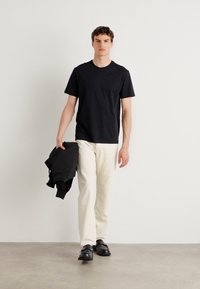 Black cotton T-shirt paired with cream trousers. Model holds a black jacket, wearing black shoes. Simple and casual design.