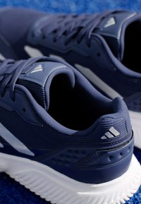 Granatowe buty sportowe z teksturowaną cholewką, białą podeszwą pośrednią i wyraźnymi akcentami logo. Posiadają sznurowany design i zaokrąglony obcas.