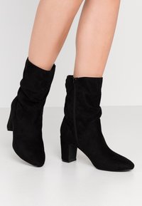 Černé semišové kotníkové boty s volným designem, špičatou špičkou a blokovým podpatkem o výšce 7 cm. Obsahují boční zip pro snadné obouvání.