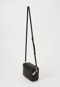 Svart rektangulär crossbodyväska med silverfärgad dragkedja och justerbar rem hängande mot en vit bakgrund.