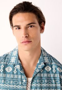 Kortærmet, blå skjorte med hvide geometriske mønstre, camp-krave og knapper, lavet af et let stof med en glat tekstur.