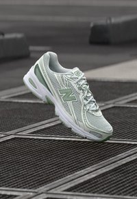 Heled roheline ja valge New Balance'i jooksujalats hõljub üle linnalike metallvõrete, taust on udune.