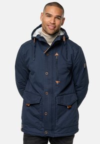 Marineblauwe geïsoleerde jas met capuchon, voorzien van een neplederen voering, leren accenten en meerdere zakken. Sluiting met knopen en rits.