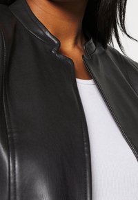 Veste en cuir noir avec un col montant, présentant une texture lisse et des détails de coutures subtils, superposée à une chemise blanche à côtes.