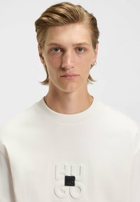 Hvid sweatshirt med præget "HUGO" tekst og et sort logo-lap på brystet. Glat overflade med rund halsudskæring.